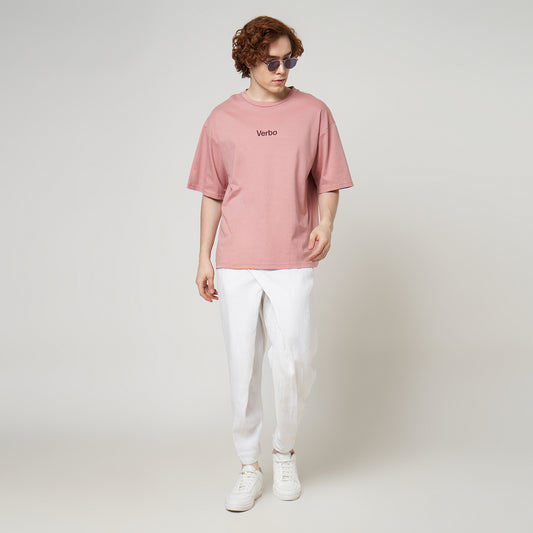 Front view of a male model wearing a rose tan boxy oversized tee with white linen trousers and white sneakers. The "Verbo" logo is visible on the chest.
