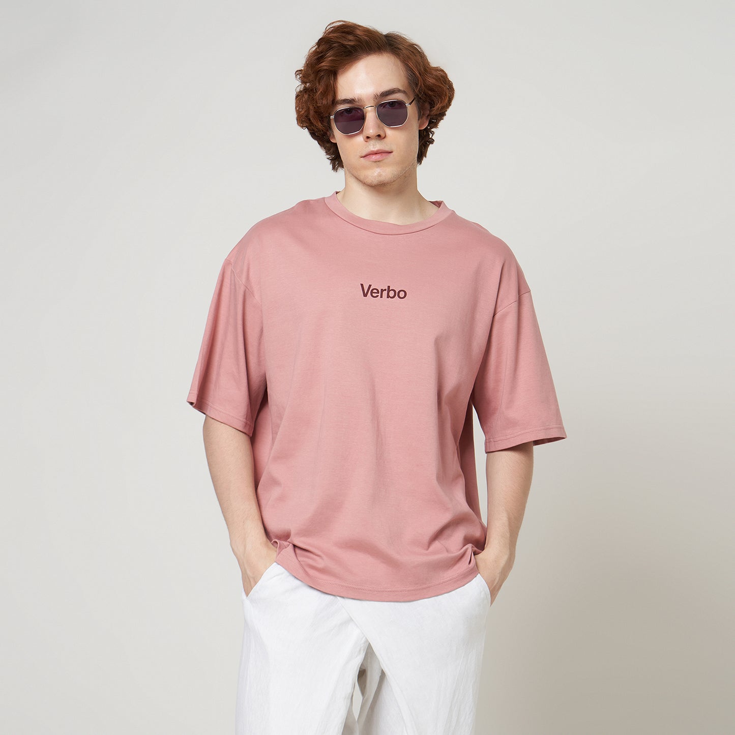 Close-up of a pink boxy oversized t-shirt featuring a small "Verbo" chest , styled with sunglasses and white pants.