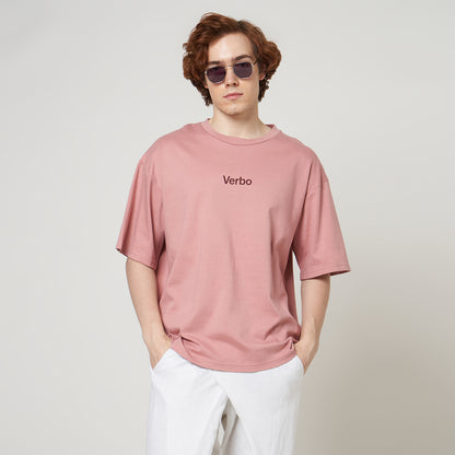 Close-up of a pink boxy oversized t-shirt featuring a small "Verbo" chest , styled with sunglasses and white pants.